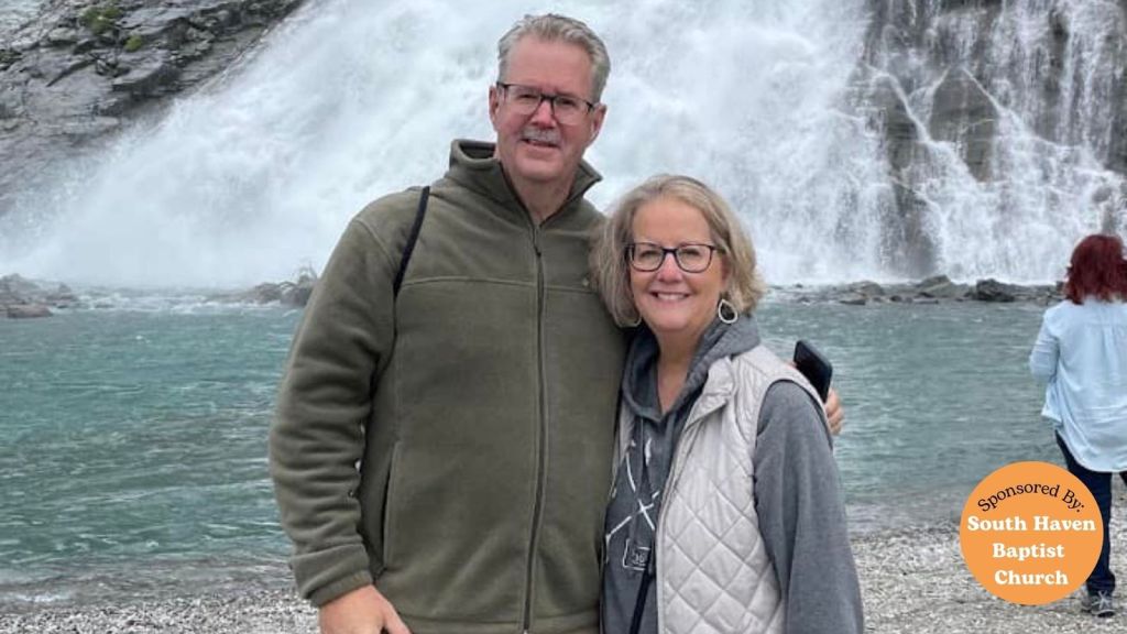 A couple stands together by a waterfall, smiling for the camera. The background features flowing water and rocky terrain.